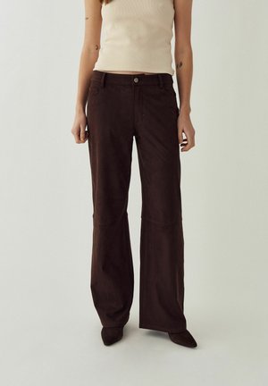 Vrouw draagt donkerbruine broek met wijde pijpen en beige mouwloos topje, staand tegen een effen lichte achtergrond.