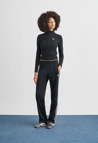 Čierna športová bunda na zips s dlhými rukávmi a skráteným strihom, doplnená čiernymi nohavicami s bielymi bočnými pruhmi a šedými športovými topánkami.