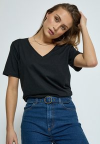 Jonge vrouw in een zwart T-shirt met V-hals en hoog uitlopende blauwe jeans met een ronde gesp, die aan haar haar zit, neutrale achtergrond.