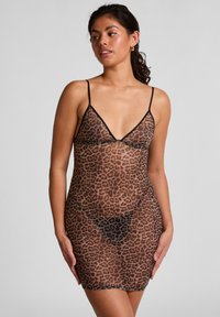 Vrouw in een doorschijnende slipdress met luipaardprint en dunne zwarte bandjes, staand tegen een effen lichtgrijze achtergrond en zijwaarts kijkend.