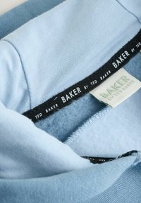 Hellblauer Kapuzenpullover aus Stoff mit weichem, strukturiertem Innenfutter. Schwarze und weiße Etiketten tragen die Aufschriften "TED BAKER" und "BAKER by TED BAKER".