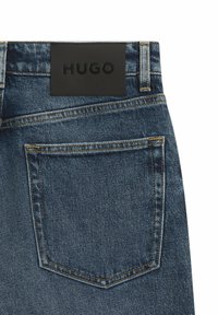 Blauwe denim jeans met een achterzak en een zwart leren label met "HUGO" erin gedrukt. De stiksels zijn in een contrasterende oranje kleur.