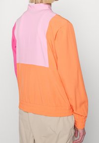 Veste à blocs de couleurs en orange, rose et rose clair. Dotée d'un col montant, d'une taille élastique et de manches longues avec poignets élastiques.