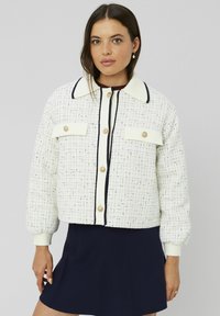 Veste blanche texturée avec des accents marine, col rond et boutons dorés. Comprend deux poches avant et des manches bouffantes.