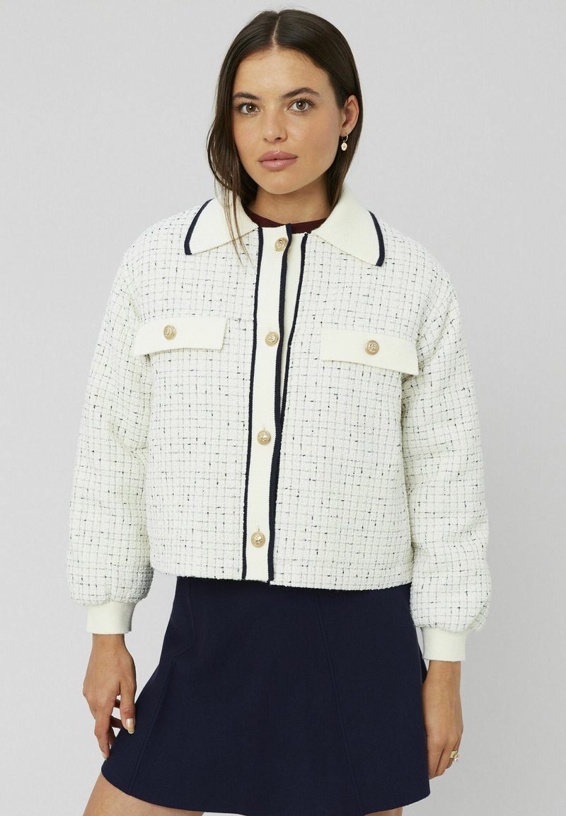 Veste blanche texturée avec des accents marine, col rond et boutons dorés. Comprend deux poches avant et des manches bouffantes.