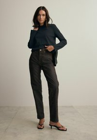 Top negro de manga larga, jeans de tiro alto color marrón oscuro con textura sutil, y sandalias de tacón negro. Diseño simple y ajustado con piernas rectas.