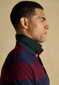 Polo krekls ar tumši zilu un vīna krāsas svītrām, kas ir ar zaļu apkakli un divām pogām. Tuvplāns uz apkakles, izcilkots uz auduma faktūru.