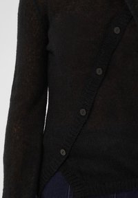 Gros plan d'une personne portant un cardigan tricoté foncé avec des boutons noirs, montrant le torse et une partie du bras gauche.