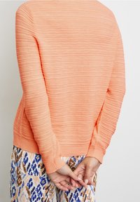 Geverfd, geribbeld oranje trui met lange mouwen, gecombineerd met gestreepte blauwe en bruine broek, handen achter de rug gekruist.