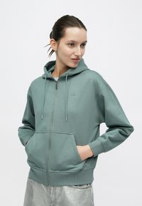 Φούτερ με φερμουάρ και κουκούλα σε πετρόλ βαμβακερό μείγμα, με μπροστινές τσέπες, ρυθμιζόμενα κορδόνια και κεντημένο λογότυπο στο στήθος.