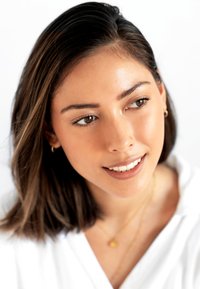 Ritratto in primo piano di una persona con capelli castano scuro fino alle spalle, che indossa una camicia bianca, una collana d'oro e piccoli orecchini a cerchio.