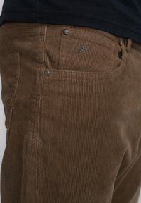 Bruine corduroy broek met verticale ribbels, voorzien van een voorzak, metalen knoop en geborduurd logo nabij de zak.