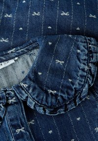 Prenda de mezclilla con un cuello con volantes, de color azul profundo, que presenta motivos de lazos blancos y detalles desgastados. El primer plano muestra la textura y la costura.