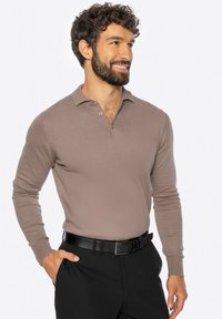Mężczyzna o kręconych ciemnych włosach i brodzie, ubrany w długi rękaw polo w kolorze taupe i czarne spodnie z czarnym paskiem, uśmiechający się i patrzący w prawo.