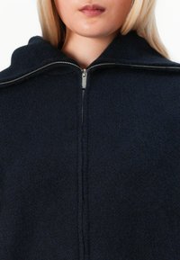 Kvinne med blondt hår som bruker en mørk marineblå zip-up fleecejakke, nærbilde fra leppene til den øvre brystkassen.