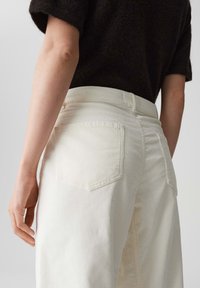 Pantaloni in corduroy bianchi con una vita alta, dotati di due tasche posteriori e una sottile rigatura verticale; abbinati a un top scuro e strutturato.