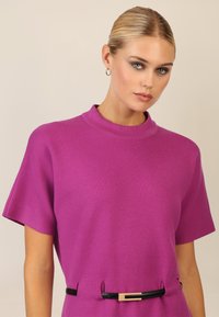 Top de punto fucsia con mangas cortas y cuello alto, que presenta un cinturón negro estrecho con una hebilla rectangular dorada en la cintura.