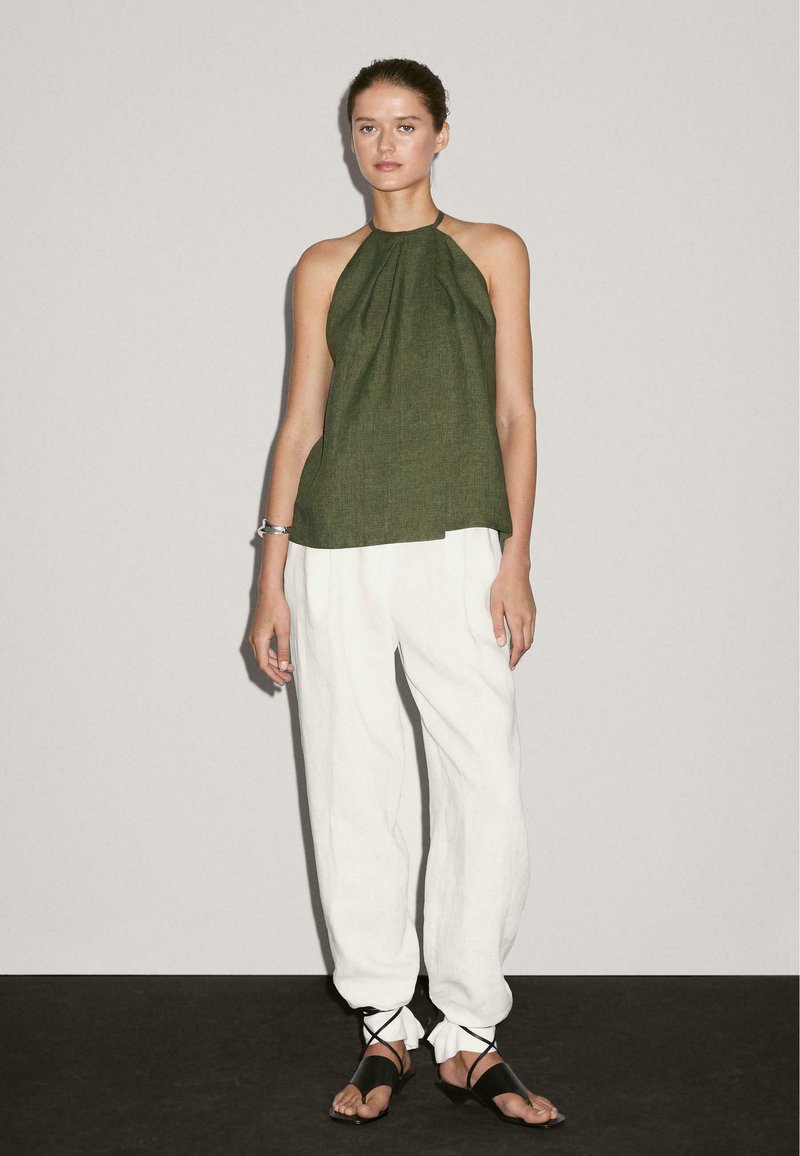 Grøn linned halterneck top parret med løse hvide linnedbukser. Sorte sandaler med ankelstropper, minimalistisk design mod en neutral baggrund.
