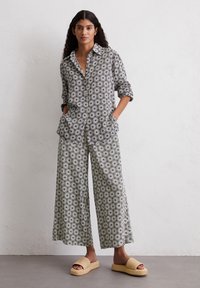 Blumenmuster, Hemd mit Knopfleiste und Kragen, hochgerollten Ärmeln, kombiniert mit weit geschnittenen Hosen. Das Model trägt sandfarbene Slipper.