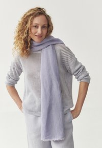 Pull en tricot gris clair à coupe ample et motif texturé, associé à une écharpe lavande douce drapée autour du cou. Ensemble décontracté.