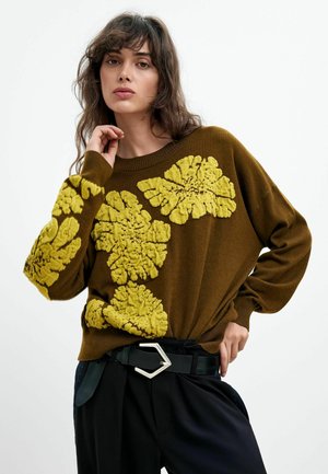 Vrouw met golvend haar draagt een bruine trui met grote gestructureerde gele bloemenpatronen en een zwarte broek met een zwarte riem met een zilveren gesp.