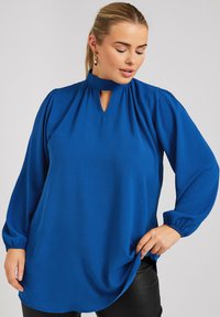 Blusa azzurra a maniche lunghe con collo alto e dettaglio a chiave, caratterizzata da spalle plissettate e maniche a polsino, realizzata in un tessuto leggero e testurizzato.