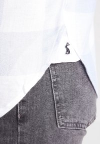 Hvidstribet skjorte med tekstureret stof, der har et logo af en kanin ved kanten. Sammen med grå denimjeans, der viser en baglomme.