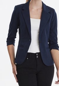 Marinblå blazer med skräddarsydd passform, slagkragar, enkel knappstängning och två framfickor, kombinerad med en vit topp och svarta byxor.