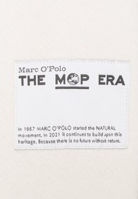 Etichetta di tessuto bianco con testo nero. "Marc O'Polo" è in cima, con "THE MOP ERA" in evidenza sotto. Il testo racconta la storia e trasmette un messaggio sulla natura.