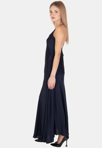 Giovane donna in piedi con un lungo vestito senza maniche blu navy, con spalline a spaghetti e tacchi alti neri, rivolta di lato.