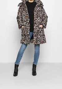 Manteau matelassé à imprimé léopard avec un col haut, une fermeture éclair et deux poches avant. Porté sur un pull noir et un jean bleu avec des bottines noires.