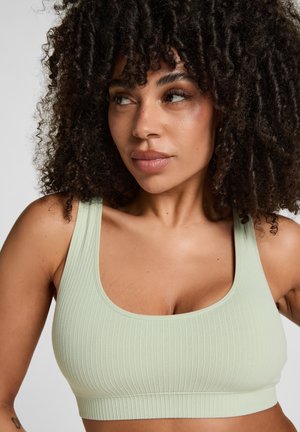 Woman with curly dark hair wearing a light green ribbed sports bra, looking to her left against a plain background.