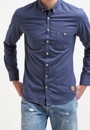 Homme portant une chemise bleue cintrée à manches longues avec une poche poitrine et un jean clair délavé, les mains posées sur les hanches.