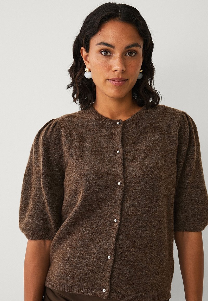 Bruine gebreide cardigan met korte pofmouwen, ronde hals en decoratieve knopen aan de voorkant, met een zachte textuur.