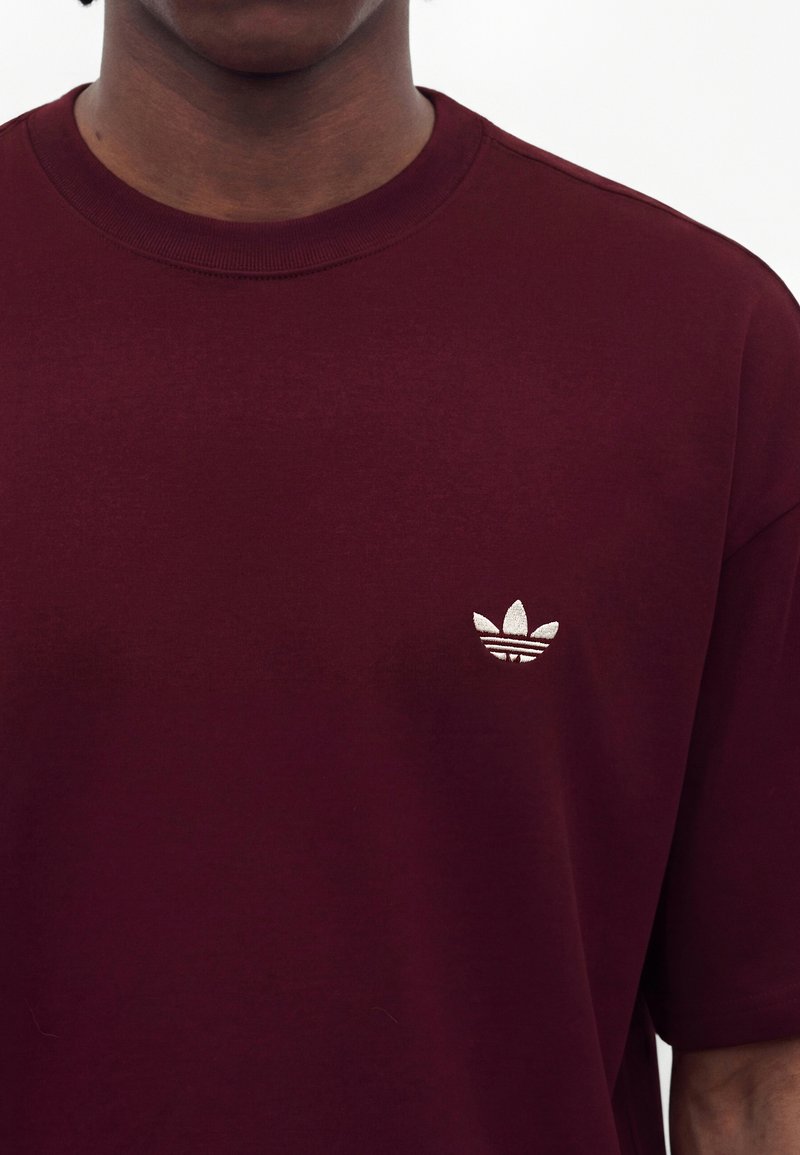 Person trägt ein dunkelrotes T-Shirt mit Rundhalsausschnitt und kleinem weißem Adidas-Trefoil-Logo auf der linken Brust.