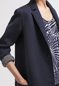 Námořnická modrá bunda se strukturovaným designem, s ohrnutými šedými manžetami, dvěma předními kapsami a hladkou texturou látky.