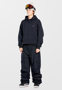 Sort hættejakke med frontlomme, sorte vandt�ætte bukser med cargo-lommer, sort-hvide sneakers, grå beanie og skibriller med orange linser.