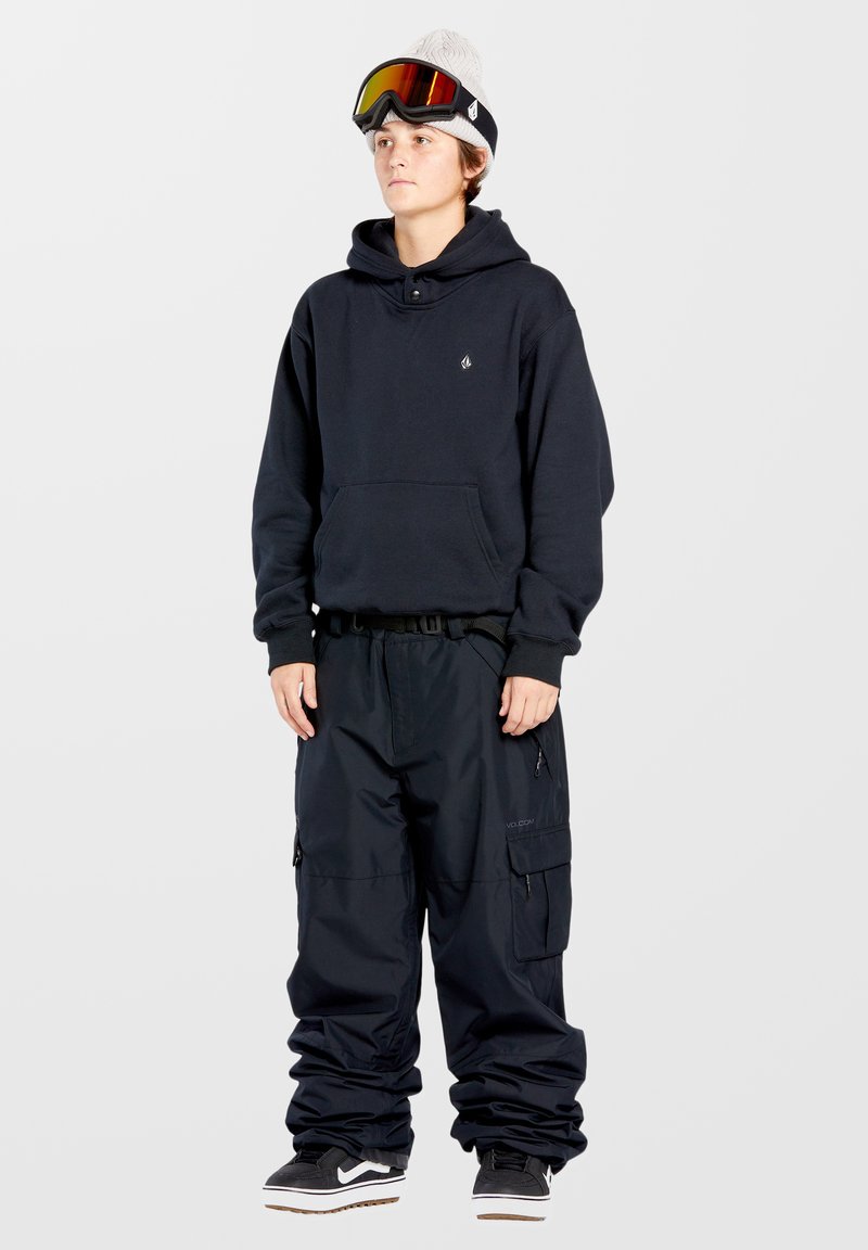 Sort hættejakke med frontlomme, sorte vandtætte bukser med cargo-lommer, sort-hvide sneakers, grå beanie og skibriller med orange linser.