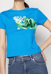 Camiseta de algodón azul con un gráfico abstracto verde y el texto "ENVI ION" en blanco. Con mangas cortas y un diseño corto.
