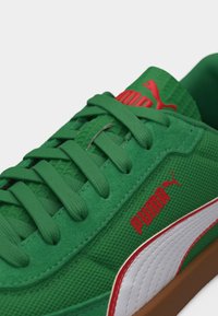 Πράσινο παπούτσι Puma με κόκκινο λογότυπο Puma, λευκή ρίγα με κόκκινο περίγραμμα, πράσινα κορδόνια και καφέ σόλα, εμφανιζόμενο σε κοντινή γωνία.