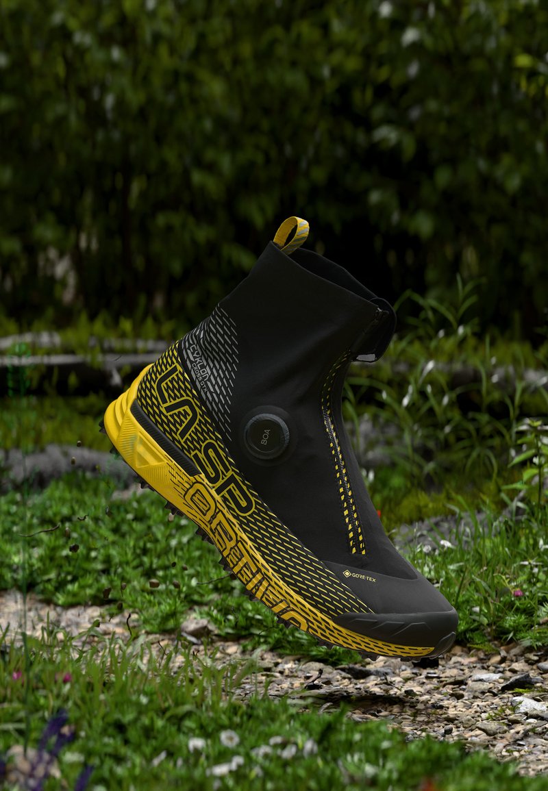 Μαύρο και κίτρινο trail running παπούτσι με ψηλή πόρπη με Gore-Tex, διακόπτη BOA και ανθεκτική σόλα, αιωρούμενο πάνω από ένα μονοπάτι από χαλίκι και γρασίδι.