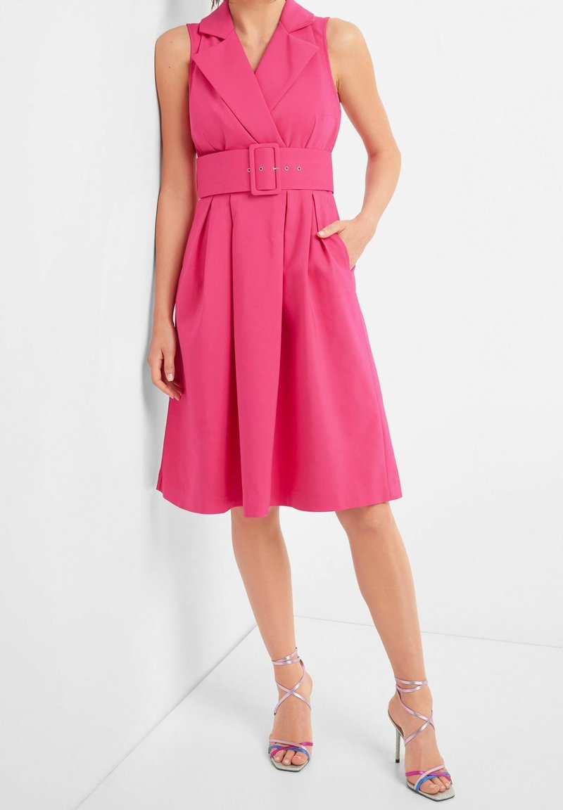 Fuchsia ärmlös klänning med bälte i midjan, skarp krage och plisserad kjol. Ackompanjeras av sandaletter med hög klack i flera färger och remmar.