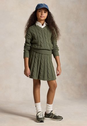 Fille aux longs cheveux bouclés portant un pull en maille torsadée vert et une jupe, une chemise blanche, une casquette bleue, des chaussettes blanches et des baskets vertes, debout sur un fond neutre.
