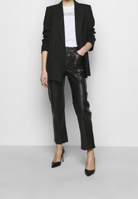 Blazer negro sobre una camiseta blanca con un logo, combinado con pantalones de cuero negros y tacones negros de punta. Textura suave y ajuste entallado.