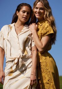Two women stand close together. One wears a cream dress with yellow and black patterns and a tie at the waist; the other wears a yellow lace dress.
