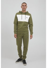 Felpa verde oliva con striscia orizzontale bianca, cappuccio con cordino e logo nero. Abbinata a pantaloni coordinati e sneakers nere.