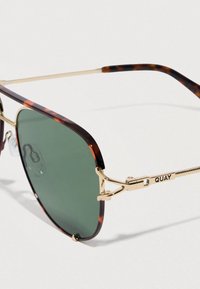 Sončna očala z okvirjem iz acetata z vzorcem želvje lupine, zelenimi lečami in zlato obarvanimi kovinskimi poudarki. Imajo nastavljive nosne blazinice in blagovno znamko QUAY.