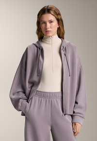 Felpa zip in un morbido viola chiaro, indossata sopra una maglia beige a collo alto. Tessuto morbido, taglio rilassato, abbinata a pantaloni coordinati.