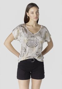 Nő mintás bézs V-nyakú felsőben és fekete öves rövidnadrágban, kezét a háta mögött pihentetve, egyszerű háttér előtt áll.