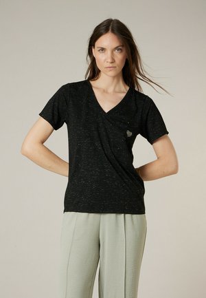 Vrouw met lang bruin haar die een zwart T-shirt met V-hals en een klein hartje draagt, gecombineerd met lichtgrijze broek, staand met handen in de zij.